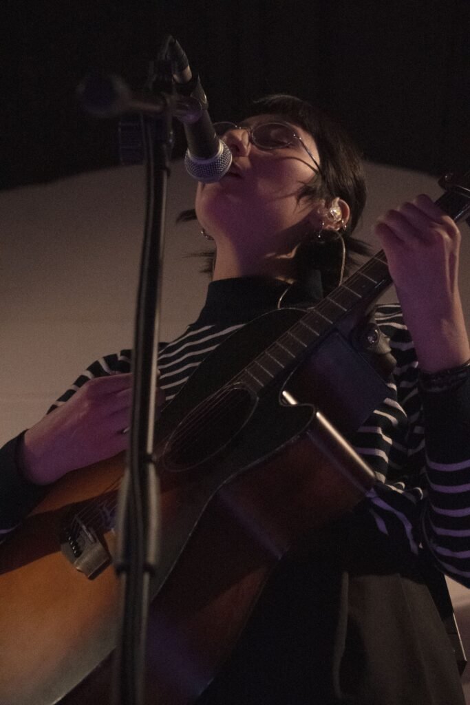 En laimagen se aprecia a la vocalista del grupo Alfilera cantando mientras toca una guitarra acustica.